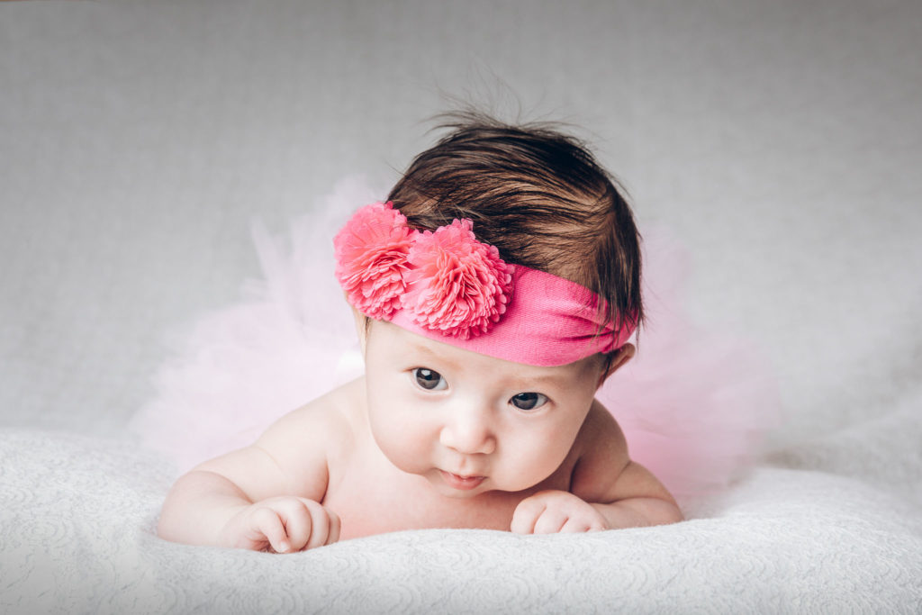 Un bébé, coiffé d'un bandeau à fleurs roses et d'une tenue en tulle blanc, est allongé sur le ventre sur une surface douce et blanche. Les yeux du bébé sont grands ouverts, semblant regarder directement l'objectif. Capturé par un talentueux photographe de famille Bordeaux, l'arrière-plan est neutre et légèrement flouté pour garder l'attention sur le bébé.