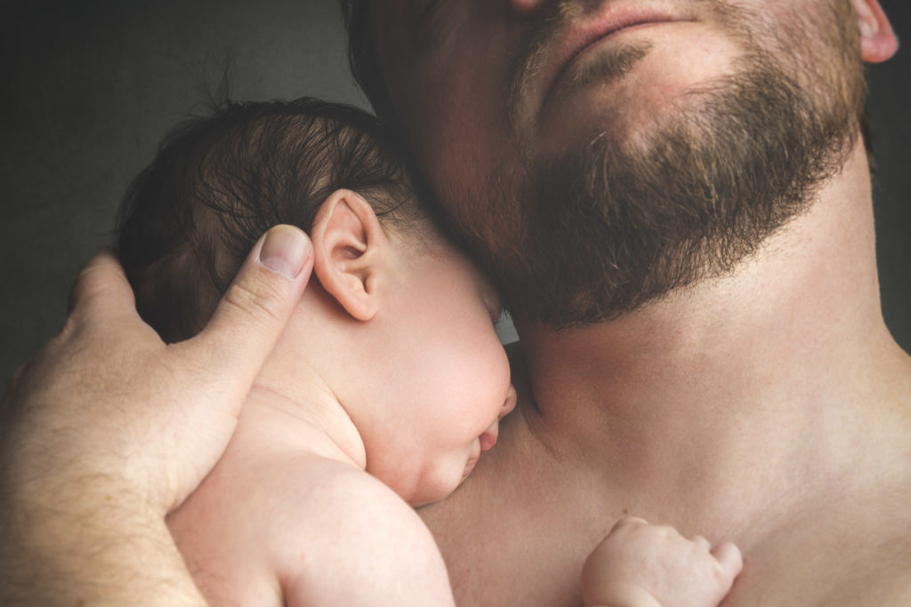 Gros plan d'un homme barbu tenant un nouveau-né endormi contre sa poitrine, capturé par un photographe de famille Bordeaux. La main de l'homme berce doucement la tête du bébé, et le petit poing de ce dernier repose sur sa poitrine. La scène chaleureuse et intime est doucement éclairée, soulignant le lien tendre qui les unit.