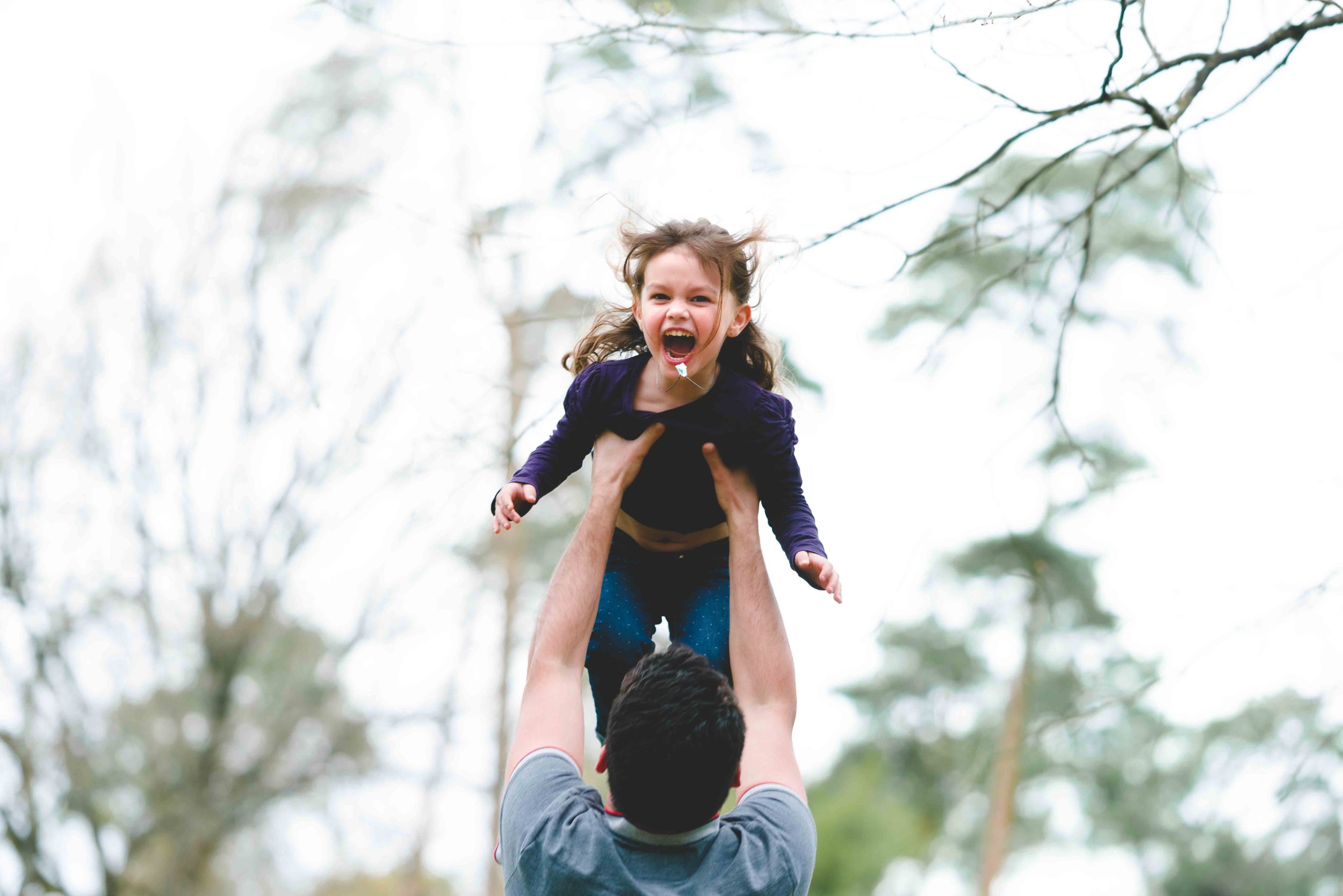 Un homme en chemise grise soulève une jeune fille aux cheveux bruns et au pull violet. La jeune fille a une expression joyeuse, la bouche ouverte et les bras écartés. Prises en photo par un photographe de famille Bordeaux, elles sont entourées de branches d'arbres nues et d'un ciel couvert.