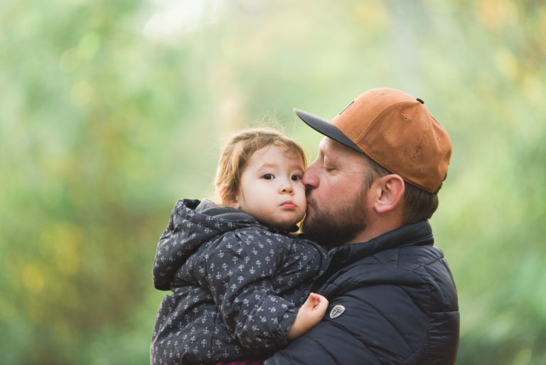 Un homme avec une casquette marron et une veste sombre tient et embrasse un jeune enfant sur la joue à l'extérieur. L'enfant, vêtu d'une veste sombre à motifs, détourne le regard de l'appareil photo pendant qu'il est tenu. L'arrière-plan est un décor naturel vert flou, suggérant un parc ou une forêt, un moment parfait capturé par photographe pour les familles.