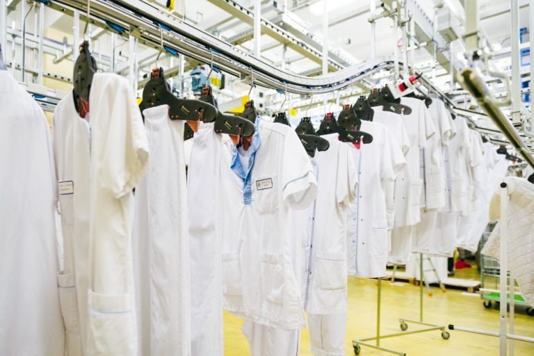 Des blouses blanches et des uniformes de travail sont suspendus sur un système de convoyeur dans un environnement industriel chez Héros Méconnus du CH d'Angoulême. Les vêtements sont soigneusement disposés en ligne et semblent fraîchement lavés. L'arrière-plan montre davantage d'équipements de blanchisserie industrielle et un environnement bien éclairé et organisé.