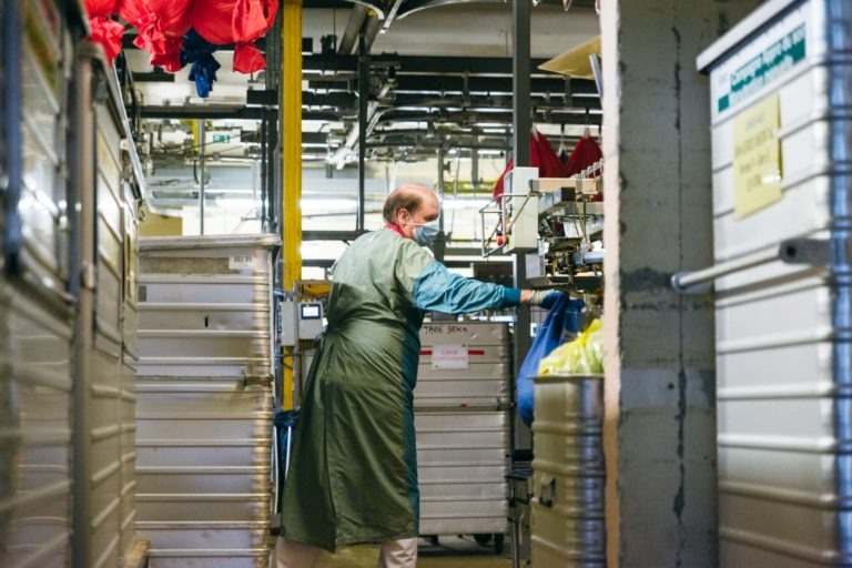 Un ouvrier portant un tablier vert, des gants bleus et un masque facial trie le linge dans une installation industrielle. Entouré de grands chariots métalliques et de bacs de rangement, il se tient debout sous des sacs à linge rouges suspendus à une rampe au-dessus. Des tuyaux et des machines industrielles forment la toile de fond de ces Héros Méconnus du CH d'Angoulême.