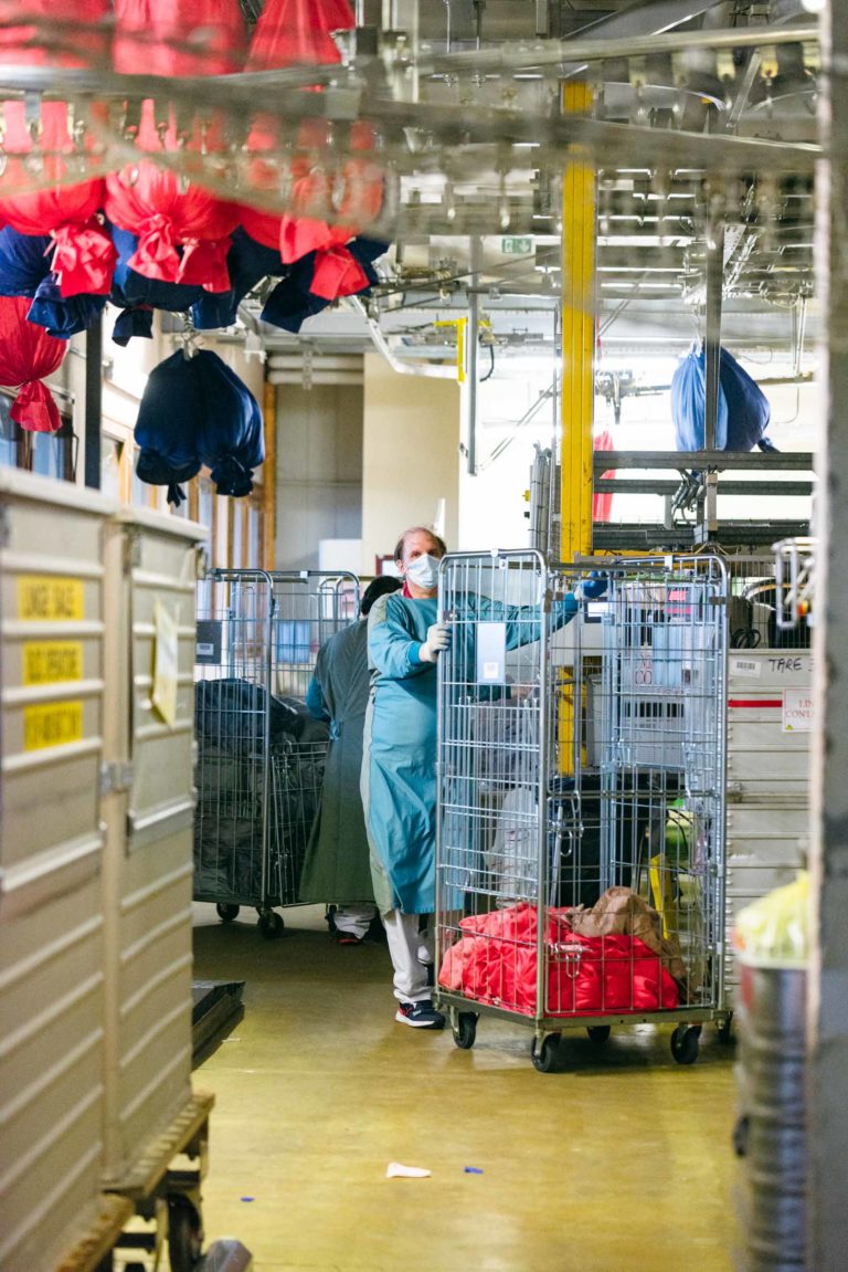 Un ouvrier portant un masque et des vêtements de protection trie des chariots à linge dans une usine. Des sacs rouges et bleus pendent du plafond et on aperçoit en arrière-plan divers équipements et cages métalliques. La scène est bien éclairée, mettant l'accent sur la propreté et l'organisation, un hommage aux Héros Méconnus du CH d'Angoulême.