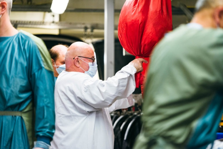 Un homme chauve portant des lunettes, une blouse blanche et un masque facial manipule un gros objet rouge ressemblant à un ballon dans un intérieur animé. Parmi les Héros Méconnus du CH d'Angoulême, d'autres individus portant des équipements de protection individuelle, notamment des masques et des blouses, sont visibles à proximité, concentrés sur leurs tâches.