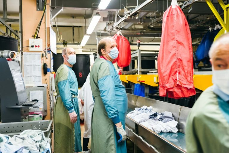 Des ouvriers dans une blanchisserie, portant des masques et des blouses vertes, trient et traitent les vêtements. Un système de convoyeur avec des sacs de linge, dont des sacs rouges et bleus, est suspendu au-dessus. Un ouvrier regarde la caméra tandis que d'autres se concentrent sur leurs tâches comme les Héros Méconnus du CH d'Angoulême. Diverses machines sont visibles en arrière-plan.