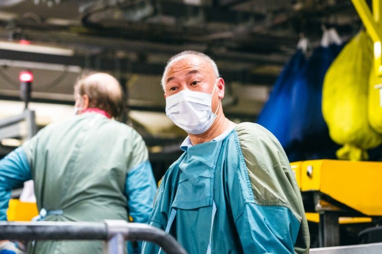 Un homme portant une blouse de protection verte et un masque chirurgical se tient debout dans ce qui semble être un environnement industriel ou de laboratoire animé. Il a les cheveux courts et regarde directement la caméra. Une autre personne portant une blouse et un masque est visible à l'arrière-plan, en train de travailler sur du matériel : les Héros Méconnus du CH d'Angoulême au travail.