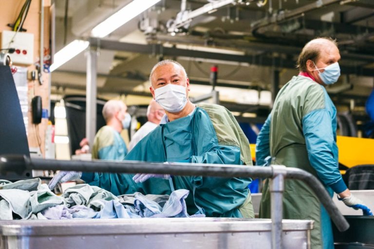 Un homme portant un masque chirurgical et une blouse verte trie des textiles dans un environnement industriel animé. Plusieurs autres travailleurs portant des vêtements similaires sont visibles à l'arrière-plan, eux aussi occupés à trier ou à faire fonctionner des machines. Ces Héros Méconnus du CH d'Angoulême semblent travailler sans relâche dans une grande blanchisserie ou une usine de transformation de textiles.