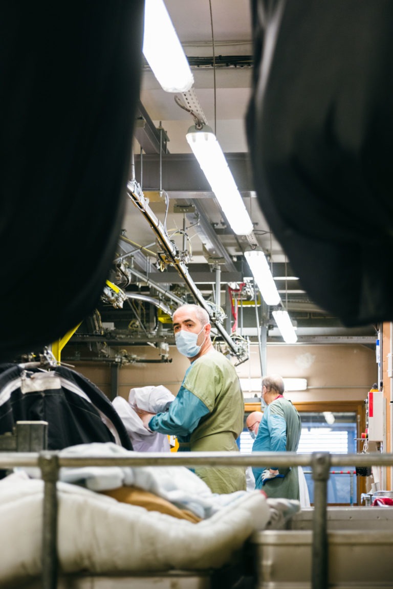 Un homme en chemise vert clair et tablier bleu porte un masque facial alors qu'il travaille dans une buanderie industrielle, incarnant l'esprit des Héros Méconnus du CH d'Angoulême. Il tient un paquet de vêtements, et derrière lui, un autre ouvrier en tenue similaire trie le linge sous des lumières fluorescentes au milieu de supports et de tuyaux métalliques.