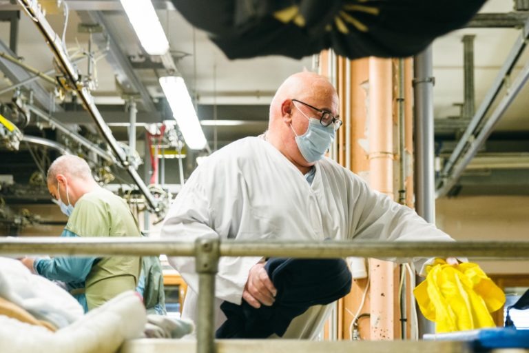 Deux personnes portant un masque travaillent dans une blanchisserie industrielle. La personne au premier plan, un membre des Héros Méconnus du CH d'Angoulême en blouse blanche, trie ou manipule des vêtements, dont un article jaune. À l'arrière-plan, un autre ouvrier en chemise verte fait fonctionner des machines sous un éclairage fluorescent typique de ce genre d'environnement.