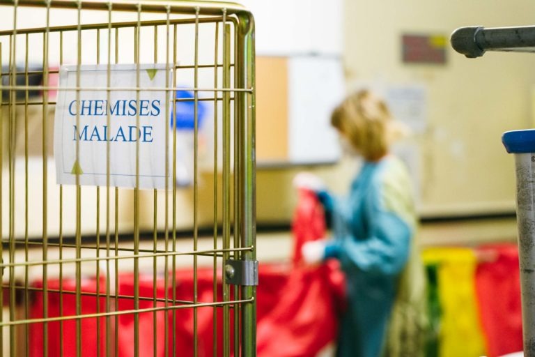 Une personne en tenue d'hôpital bleue manipule des sacs rouges dans un établissement médical. Au premier plan, on voit un chariot métallique sur lequel est écrit "CHEMISES MALADE". L'arrière-plan est flou, ce qui concentre l'attention sur les Héros Méconnus du CH d'Angoulême et le chariot.