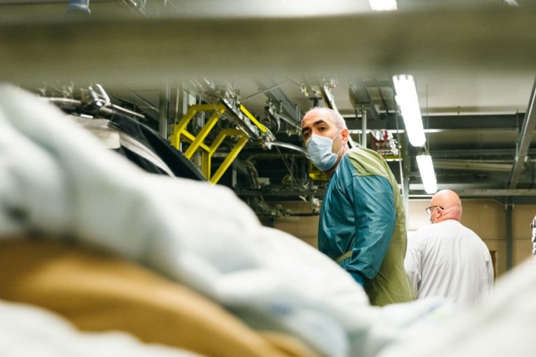 Deux personnes travaillent dans une usine de transformation de linge ou de textiles. Un travailleur au premier plan porte un équipement de protection, notamment une blouse bleue et un masque, et se tourne vers la caméra. La deuxième personne, tournée vers l'extérieur, porte une blouse blanche. L'arrière-plan montre de grandes machines et un éclairage intense. Héros Méconnus du CH d'Angoulême!