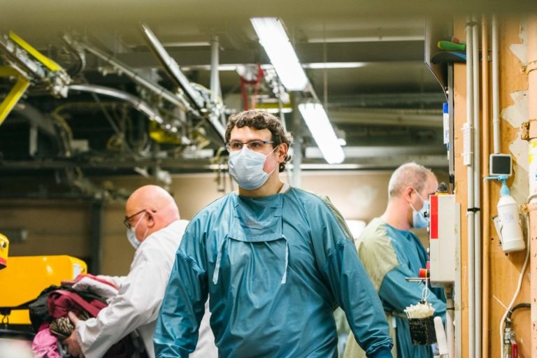 Trois professionnels de la santé, tous portant des masques et des blouses de protection, sont vus en train de travailler dans un établissement médical doté d'un éclairage industriel. Le personnage central, drapé de bleu sarcelle, regarde vers l'avant tandis que deux collègues, l'un tenant des objets, s'occupent de tâches en arrière-plan au milieu de divers équipements. Ce sont les Héros Méconnus du CH d'Angoulême.