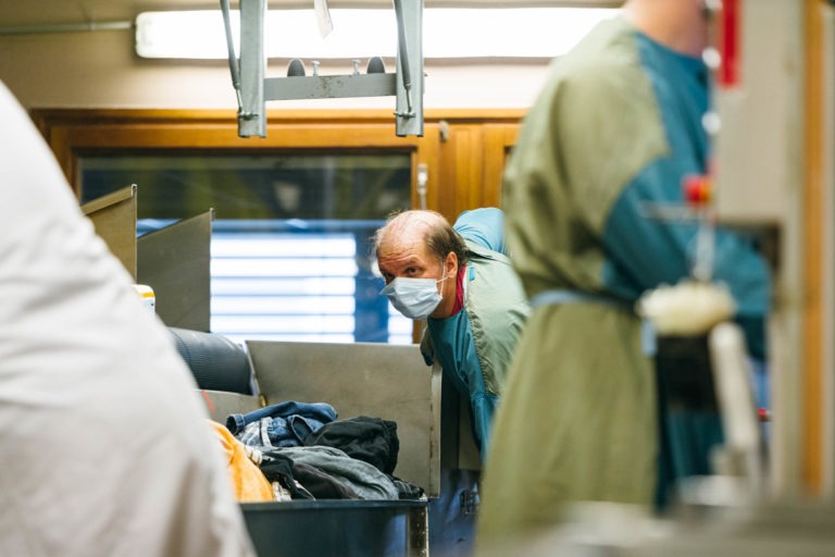 Une personne portant un masque et une tenue médicale est penchée sur un comptoir en acier inoxydable, le regard fixé devant elle. Au premier plan, un autre individu portant une tenue similaire est partiellement visible. Le décor semble être celui d'un environnement médical ou de laboratoire avec divers équipements et supports à proximité - vraiment les Héros Méconnus du CH d'Angoulême.