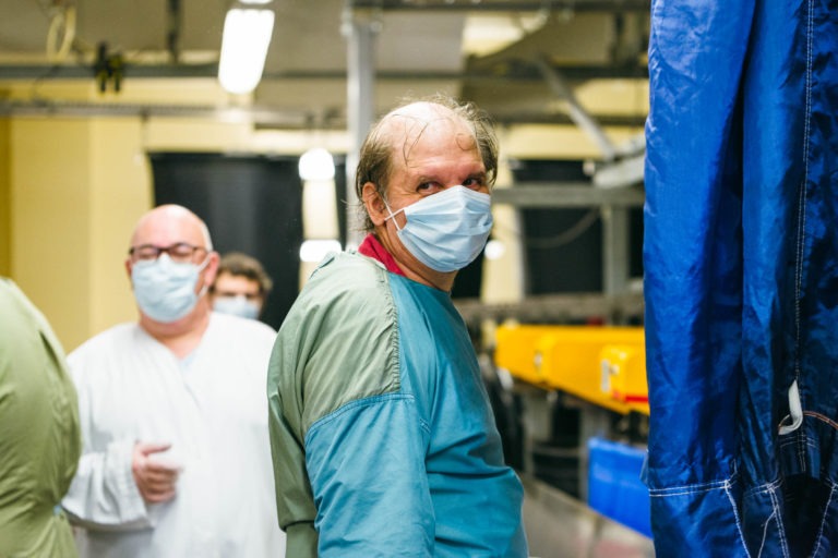 Un homme porte un masque et une blouse de protection bleue et regarde la caméra dans un décor de laboratoire. Un autre homme masqué et vêtu de blanc se tient à proximité. Derrière eux, des étagères et du matériel témoignent d'un environnement professionnel axé sur la sécurité et l'hygiène, digne des Héros Méconnus du CH d'Angoulême.