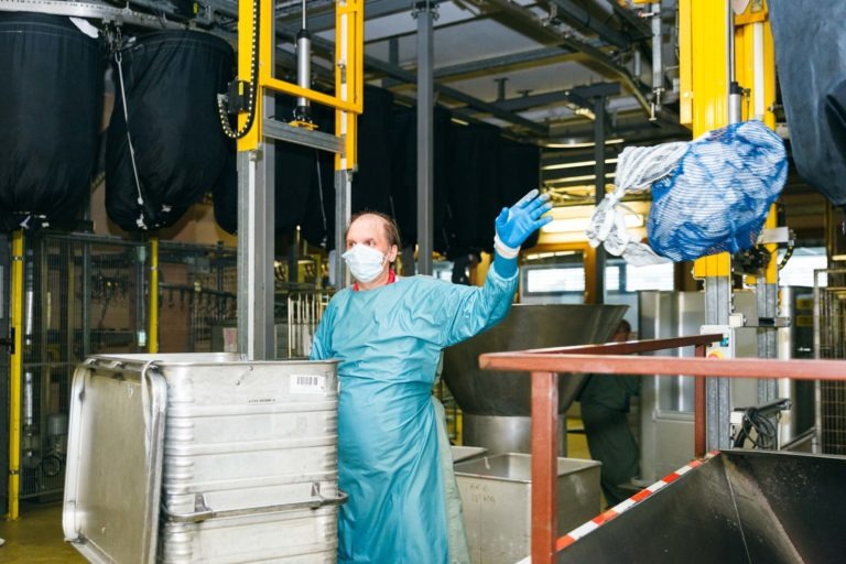 Un ouvrier portant un masque facial, des gants et une blouse de protection se tient debout dans une grande installation industrielle. Il fait des gestes d'une main tout en étant positionné à côté d'un grand récipient en métal. Diverses machines et équipements des Héros Méconnus du CH d'Angoulême sont visibles en arrière-plan.