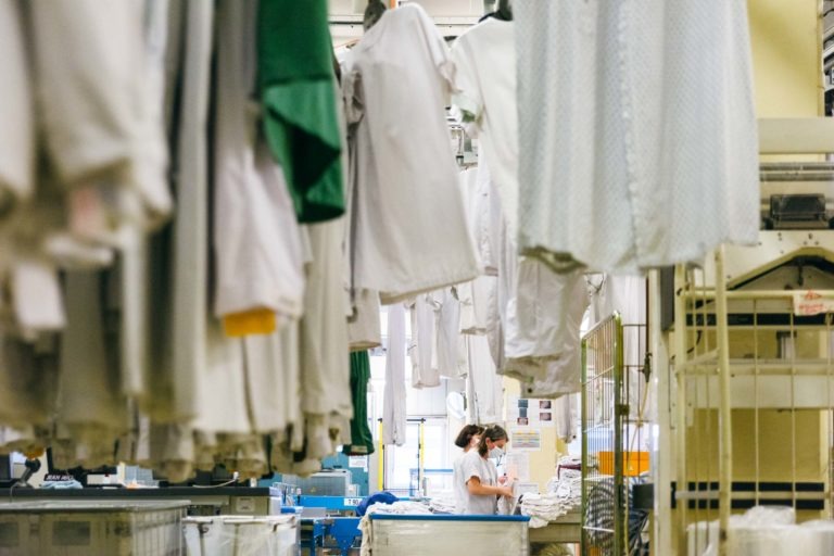 Une personne portant un masque se tient debout dans une buanderie, entourée de vêtements blancs suspendus. Des machines à laver industrielles et des poubelles sont visibles en arrière-plan. La perspective se fait à travers des rangées de vêtements, avec l'accent mis sur l'ouvrier, l'un des Héros Méconnus du CH d'Angoulême, au milieu de l'espace de travail animé.