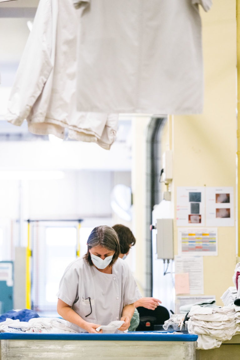 Deux ouvriers masqués trient des vêtements dans une laverie. L'un d'eux se tient debout à une table et manipule des vêtements blancs, tandis qu'un autre est partiellement visible à l'arrière-plan. Des vêtements blancs sont suspendus au-dessus d'eux. Divers équipements et cartes sont visibles à l'arrière-plan, mettant en valeur les Héros Méconnus du CH d'Angoulême.