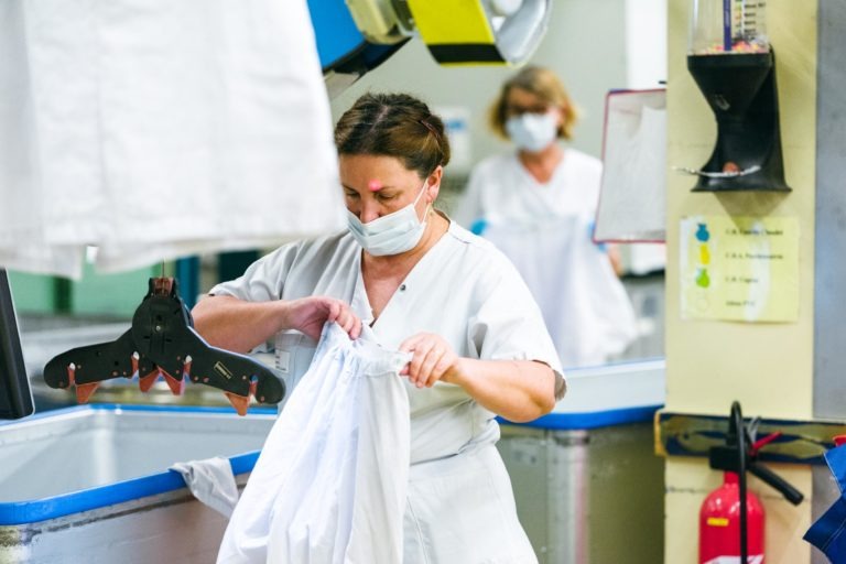 Deux soignants portant des uniformes blancs et des masques de protection travaillent dans une buanderie. Un travailleur au premier plan plie un drap blanc près d'un vêtement suspendu avec des pinces à linge, incarnant l'esprit des Héros Méconnus du CH d'Angoulême. Le deuxième travailleur à l'arrière-plan manipule également du linge au milieu de divers bacs et équipements.