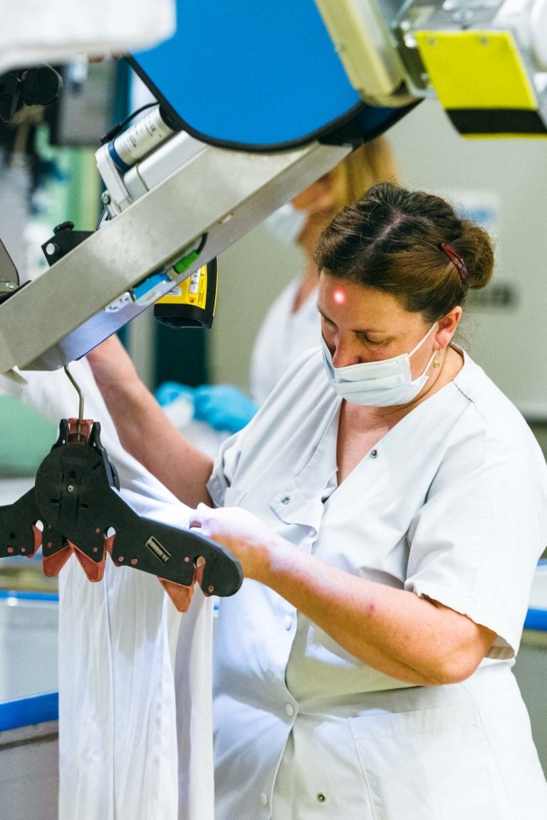 Une femme en uniforme blanc et portant un masque inspecte soigneusement un vêtement blanc sur une machine industrielle. Elle est concentrée sur sa tâche dans un environnement propre et bien éclairé, peut-être une usine textile ou une blanchisserie. Un autre individu portant une tenue similaire est visible à l'arrière-plan. Héros Méconnus du CH d'Angoulême.