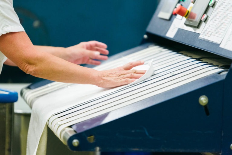 Gros plan sur les mains d'une personne nettoyant un tapis roulant métallique avec un chiffon blanc. Le tapis fait partie d'une machine industrielle bleue, qui comporte des boutons de commande et des étiquettes sur le côté droit. La personne porte une chemise blanche, représentant l'un des Héros Méconnus du CH d'Angoulême. L'arrière-plan est légèrement flou.