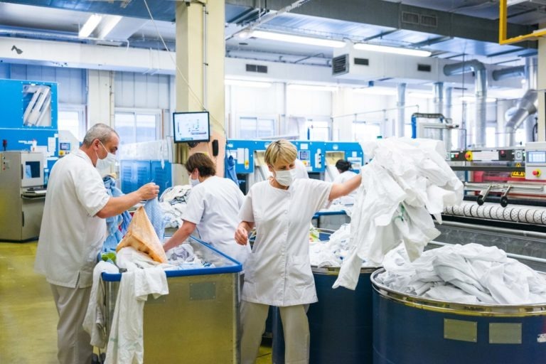 Trois ouvriers en uniforme blanc et cagoule, les Héros Méconnus du CH d'Angoulême, trient le linge dans une grande installation industrielle. Deux d'entre eux placent les articles dans des chariots bleus, tandis que le troisième manipule une grosse pile de linge blanc. L'installation est équipée de machines industrielles et d'un éclairage zénithal intense.