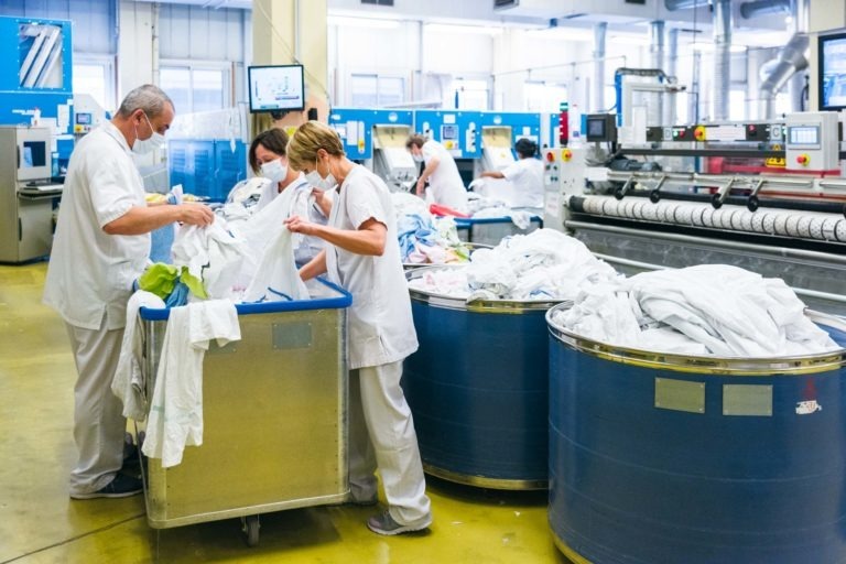 Des ouvriers en uniformes blancs et masqués, les Héros Méconnus du CH d'Angoulême, trient le linge dans une grande et lumineuse installation industrielle. Plusieurs chariots bleus débordent de linge et de vêtements. Des machines et des écrans sont visibles en arrière-plan, indiquant un environnement hautement automatisé. La scène est animée et organisée.