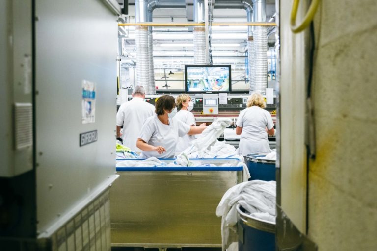 Des ouvriers en uniforme blanc trient du linge blanc autour de grands bacs en métal dans une blanchisserie industrielle. Le décor est composé d'équipements en acier inoxydable, de conduits et d'écrans muraux affichant les détails du traitement du linge. L'environnement semble propre et organisé, à l'image des Héros Méconnus du CH d'Angoulême.