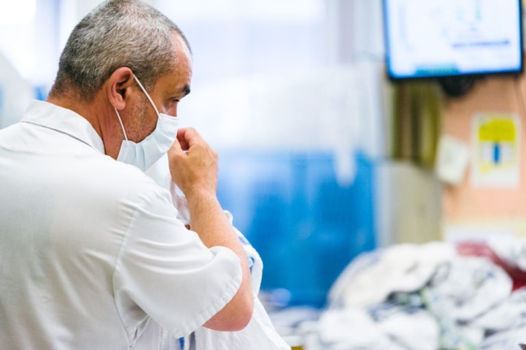 Un soignant en uniforme blanc ajuste un masque facial debout dans un hôpital ou un milieu médical. Divers équipements et fournitures médicales sont visibles en arrière-plan, ainsi qu'un écran de télévision affichant des informations floues. Véritable Héros Méconnus du CH d'Angoulême, le soignant est concentré sur la tâche, faisant preuve de dévouement et d'attention.