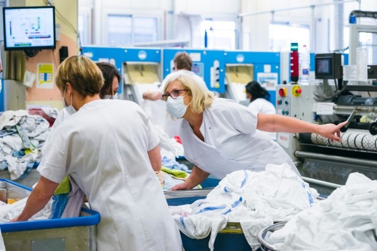 Plusieurs ouvriers en uniforme blanc et masqués trient et inspectent le linge dans une blanchisserie industrielle. L'espace de travail est rempli de grands bacs bleus contenant du linge, et diverses machines industrielles sont visibles en arrière-plan. Ces Héros Méconnus du CH d'Angoulême veillent aux normes d'hygiène pour tous.