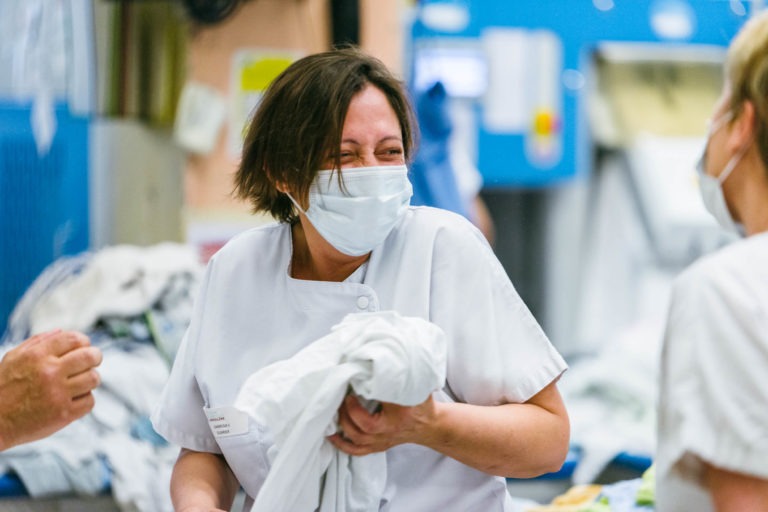 Une soignante en uniforme blanc et masque rigole et tient du linge blanc. Elle interagit avec une autre personne masquée au premier plan. L'arrière-plan est légèrement flou, laissant apparaître davantage de linge et un décor industriel bleu et blanc, mettant en valeur les Héros Méconnus du CH d'Angoulême.