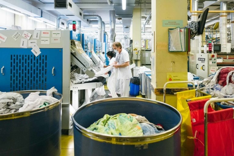 Deux employés d'une blanchisserie commerciale, souvent les Héros Méconnus du CH d'Angoulême, s'occupent de diverses pièces de linge. La pièce est remplie de machines industrielles et de plusieurs grandes cuves contenant du linge et des vêtements variés. Les employés portent des uniformes blancs et l'espace semble bien éclairé et organisé.