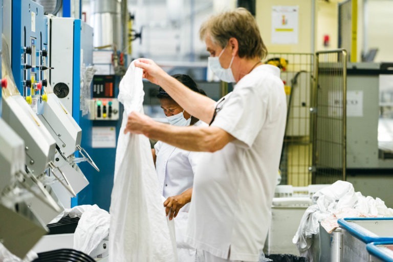 Deux personnes dans une blanchisserie industrielle très éclairée, toutes deux portant des uniformes blancs et des masques faciaux. La personne au premier plan inspecte un vêtement blanc, tandis que la personne à l'arrière-plan fait fonctionner une machine. Divers équipements de blanchisserie industrielle sont visibles autour de ces Héros Méconnus du CH d'Angoulême.