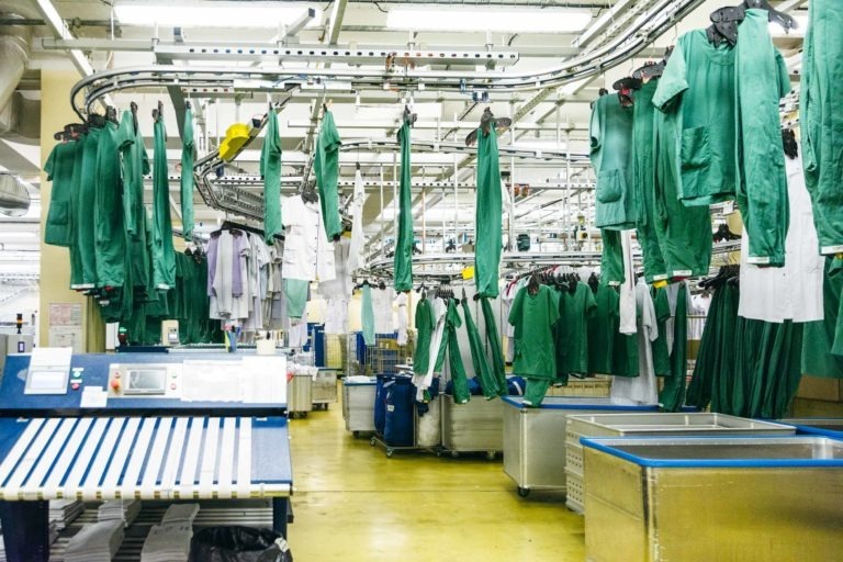 Une buanderie d'hôpital avec de nombreux uniformes verts et blancs suspendus à un système de convoyeur. Le sol est jaune et l'espace est rempli de grands bacs à linge bleus et argentés. Des postes de travail avec des panneaux de contrôle sont visibles au premier plan, mettant en valeur un décor industriel où les Héros Méconnus du CH d'Angoulême travaillent sans relâche.