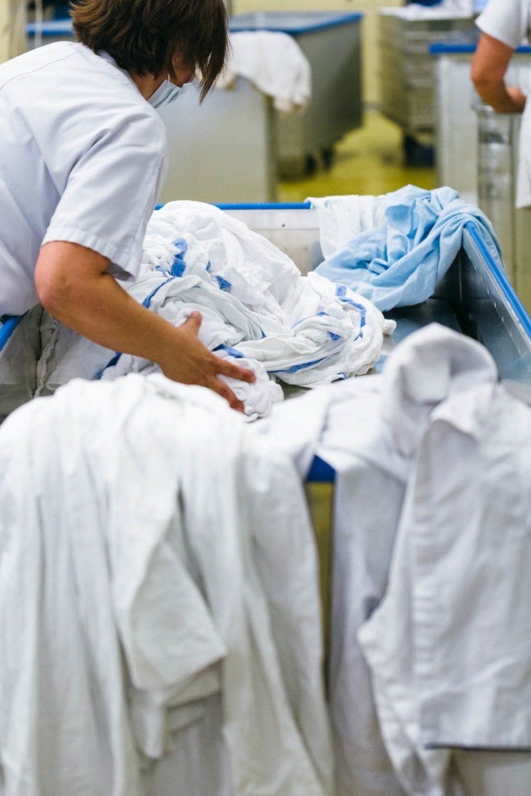 Un personnage manipule une grande pile de textiles blancs et bleu clair, probablement du linge, sur un convoyeur horizontal. L'individu, l'un des Héros Méconnus du CH d'Angoulême, est vêtu d'un uniforme blanc et vu de dos, son visage n'étant pas visible. Le décor semble être celui d'une blanchisserie ou d'une usine de traitement de textiles.