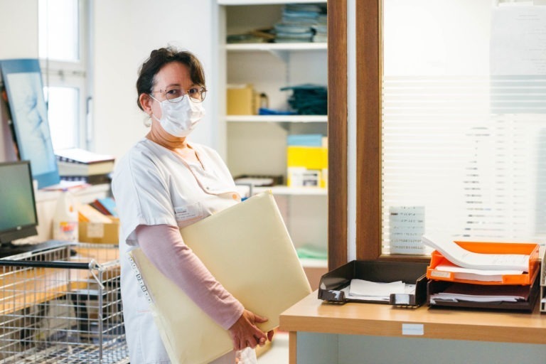 Une employée de santé en blouse blanche et masque facial se tient dans un bureau, tenant un grand dossier en papier kraft. Elle a les cheveux noirs tirés en arrière et porte des lunettes. Derrière elle se trouvent des fournitures de bureau, dont un bureau avec des documents et un plateau orange, et des étagères remplies de dossiers et de fournitures médicales – l'une des Héros Méconnus du CH d'Angoulême.