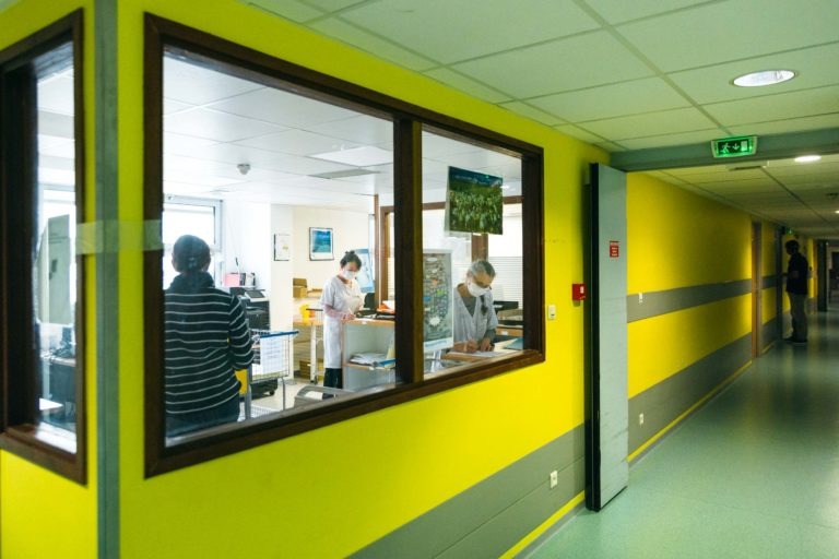 Vue à travers une fenêtre d'un bureau d'hôpital où trois professionnels de la santé portant des masques et des blouses travaillent avec diligence. Le couloir extérieur, aux murs jaunes et aux accents gris, montre une personne seule en train de marcher. La scène est bien éclairée par des lampes fluorescentes suspendues, capturant les Héros Méconnus du CH d'Angoulême en action.