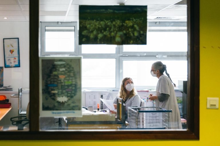 Une photographie prise à travers une fenêtre montre deux professionnels de la santé en conversation, incarnant l'esprit des Héros Méconnus du CH d'Angoulême. Tous deux portent des blouses blanches et des masques faciaux. L'une est assise à un bureau avec des papiers tandis que l'autre se tient à côté d'elle. La pièce est lumineuse, avec de grandes fenêtres et des affiches sur les murs.