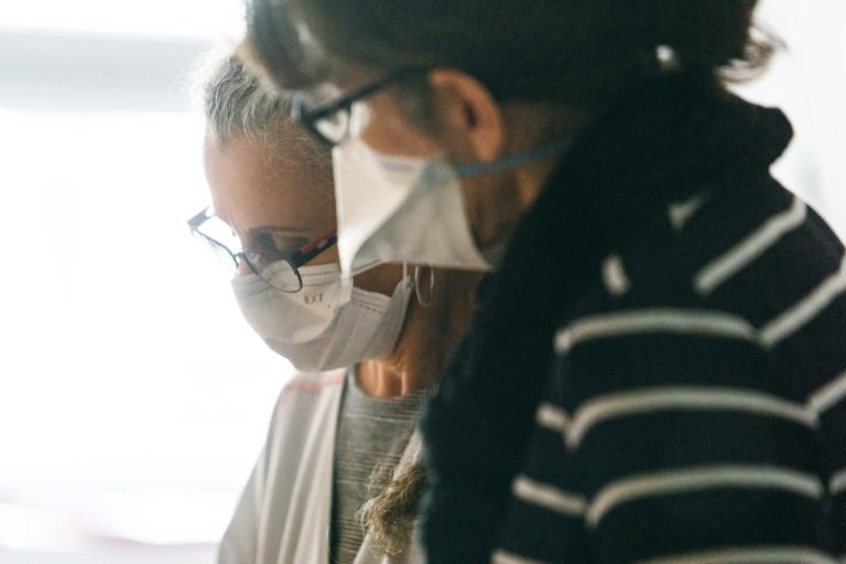 Deux individus portant des masques et des lunettes sont vus en consultation. La personne de gauche, revêtue d'une blouse de laboratoire, fait allusion à un environnement de soins de santé. Ils semblent absorbés par une discussion, concentrés sur une tâche ou un document devant eux - de véritables Héros Méconnus du CH d'Angoulême.