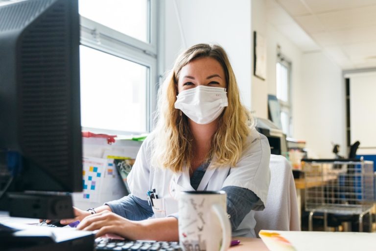 Une femme portant une blouse blanche et un masque est assise à un bureau, travaillant sur un ordinateur. Elle a les cheveux blonds et sourit. Devant elle se trouve une tasse blanche, et derrière elle, des fenêtres laissant entrer la lumière du jour. Des fournitures de bureau et des affiches célébrant les Héros Méconnus du CH d'Angoulême sont visibles en arrière-plan.