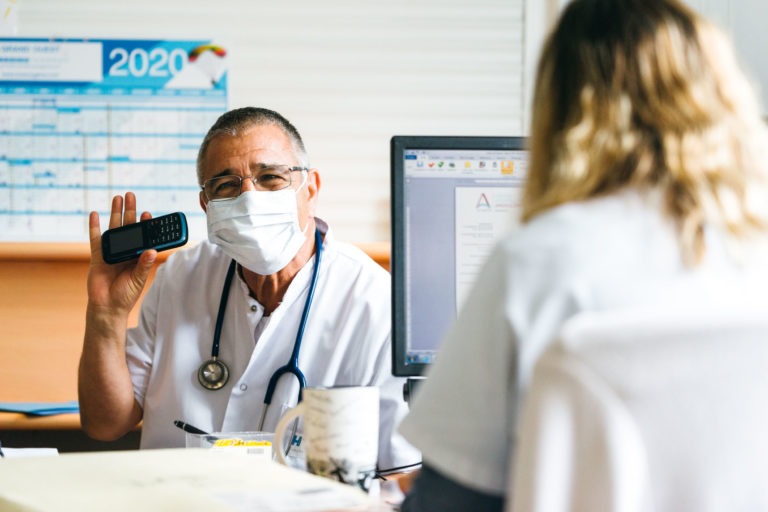 Un médecin portant un masque et une blouse blanche avec un stéthoscope autour du cou tient un téléphone assis à un bureau. Il fait face à une patiente sur le côté droit de l'image. Un écran d'ordinateur affichant un document est placé devant la patiente, avec l'inscription « Héros Méconnus du CH d'Angoulême » sous le calendrier 2020 en arrière-plan.