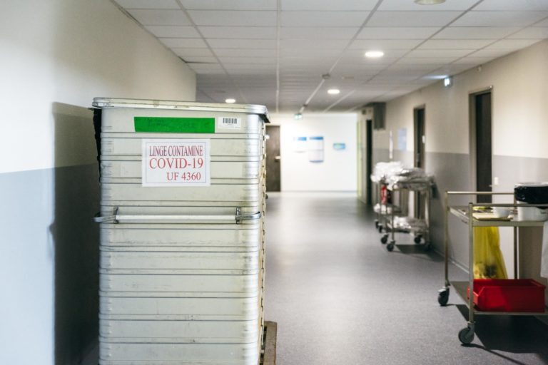 Un couloir d'hôpital avec un grand conteneur en métal étiqueté « LINGE CONTAMINE COVID-19 UF 4360 » bien visible sur la gauche. Le couloir est vide, avec des chariots et des fournitures médicales le long des murs. L'éclairage est vif et le couloir semble propre, un témoignage méconnu des Héros Méconnus du CH d'Angoulême.