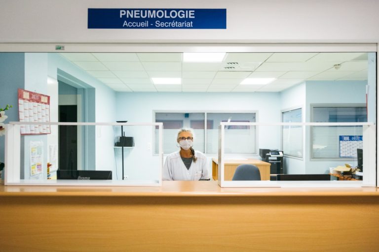 Un professionnel de la santé, l'un des Héros Méconnus du CH d'Angoulême, porte un masque et une blouse blanche alors qu'il se tient derrière un bureau d'accueil dans une clinique de pneumologie. Le bureau est doté de parois en verre et un panneau bleu au-dessus indique « PNEUMOLOGIE Accueil - Secrétariat ». Le mobilier de bureau et le matériel médical remplissent l'arrière-plan.