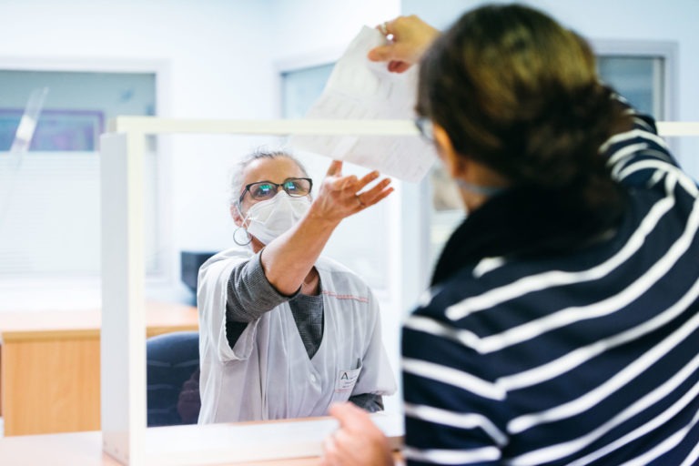 Un soignant masqué, membre des Héros Méconnus du CH d'Angoulême, assis derrière un écran de protection transparent, tend la main pour prendre un document des mains d'une personne portant des lunettes et une chemise rayée. Le décor semble être celui d'un cabinet médical ou administratif avec des bureaux et des fenêtres en arrière-plan.