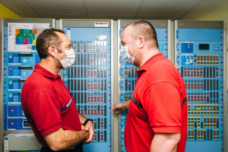 Deux hommes en chemise rouge et masque facial se tiennent face à face, en pleine conversation devant des racks de serveurs informatiques bleus dotés de nombreux boutons et interfaces dans une salle de contrôle. L'homme de gauche a les bras croisés, tandis que l'homme de droite fait un geste de la main droite. Ces Héros Méconnus du CH d'Angoulême travaillent sans relâche dans les coulisses.