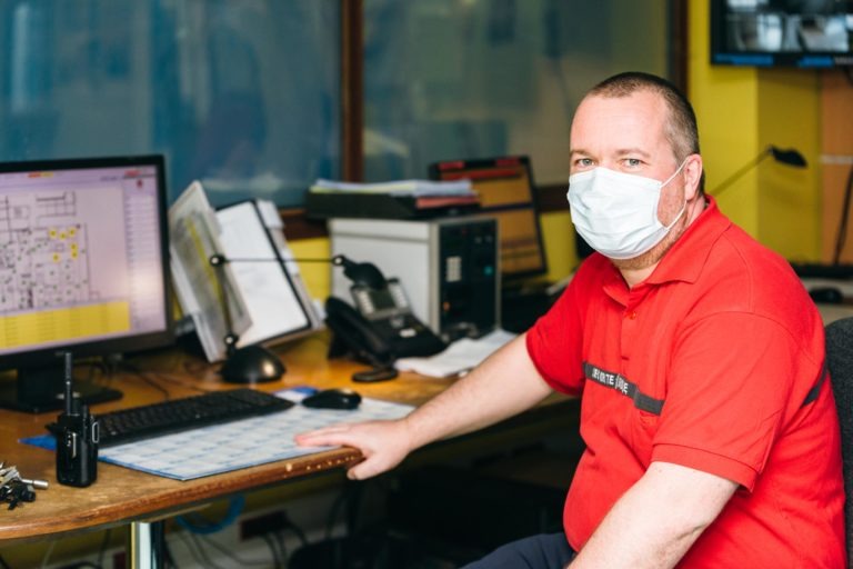 Un homme portant un masque facial est assis à un bureau dans un environnement de bureau, incarnant l'esprit des Héros Méconnus du CH d'Angoulême. Il est vêtu d'un polo rouge et a la main posée sur le bureau où sont placés un ordinateur, un clavier et d'autres équipements de bureau. Une carte ou un plan est affiché sur l'écran, entouré de papiers et de classeurs.