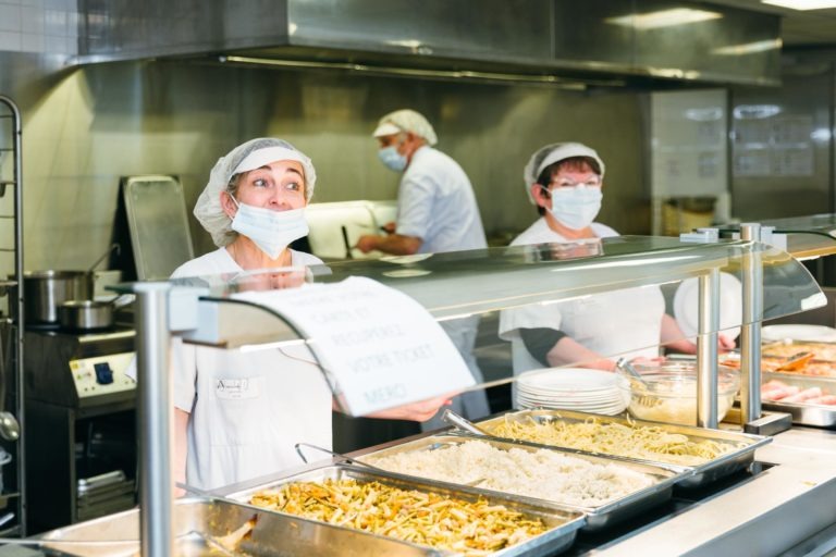 Deux employés de cafétéria, Héros Méconnus du CH d'Angoulême, portent des uniformes blancs, des filets à cheveux et des masques alors qu'ils se tiennent derrière un comptoir de service dans une cuisine professionnelle. Ils servent des plateaux de nourriture, notamment du riz et des nouilles, à des clients invisibles. Une cuisine industrielle équipée d'équipements en acier inoxydable est visible en arrière-plan.