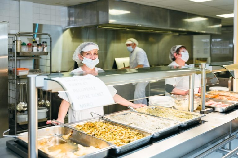 Trois employés de la cafétéria, les Héros Méconnus du CH d'Angoulême, portent des masques, des filets à cheveux et des uniformes blancs alors qu'ils servent de la nourriture derrière un comptoir rempli de plateaux de pâtes et de riz. Une vitre en plexiglas les sépare des clients. Une pancarte sur le comptoir demande le ticket et remercie les clients.