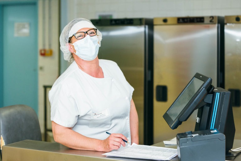 Une professionnelle de la santé en uniforme blanc, masque et filet à cheveux se tient devant un comptoir avec un bloc-notes et un stylo. Elle regarde devant elle, derrière un système informatique noir et bleu, avec des équipements industriels en acier inoxydable en arrière-plan. Le décor semble être celui d'un hôpital ou d'un établissement médical, un véritable Héros Méconnu du CH d'Angoulême.