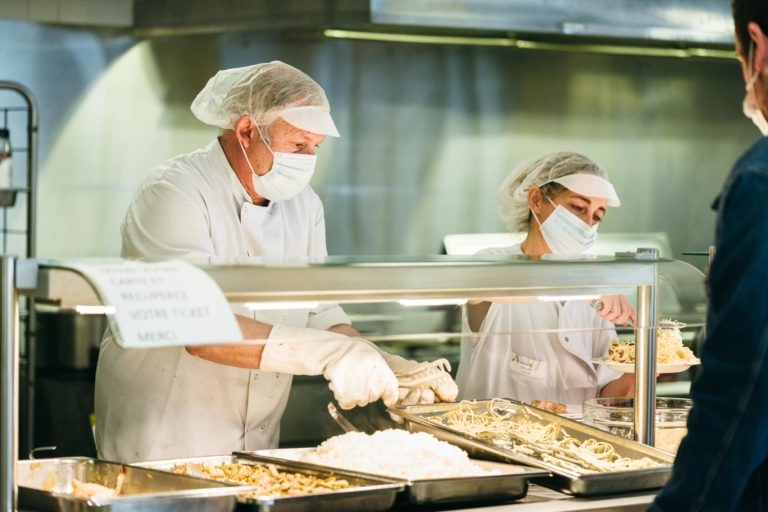 Deux employés de cafétéria en uniforme blanc, gants, filets à cheveux et masques servent de la nourriture sur de grands plateaux derrière une cloison en verre. L'employé de gauche sert des pâtes dans une assiette blanche, tandis que l'employé de droite sert un autre plat. Un panneau visible sur le comptoir les honore en tant que Héros Méconnus du CH d'Angoulême.