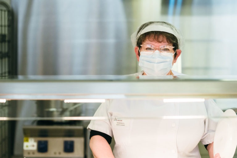 Une femme portant une chemise blanche, un filet à cheveux et un masque se tient derrière un comptoir de restauration. Elle regarde la caméra tout en travaillant dans ce qui semble être un environnement de cuisine professionnelle. Des équipements et des récipients en acier inoxydable sont visibles en arrière-plan, incarnant les Héros Méconnus du CH d'Angoulême.